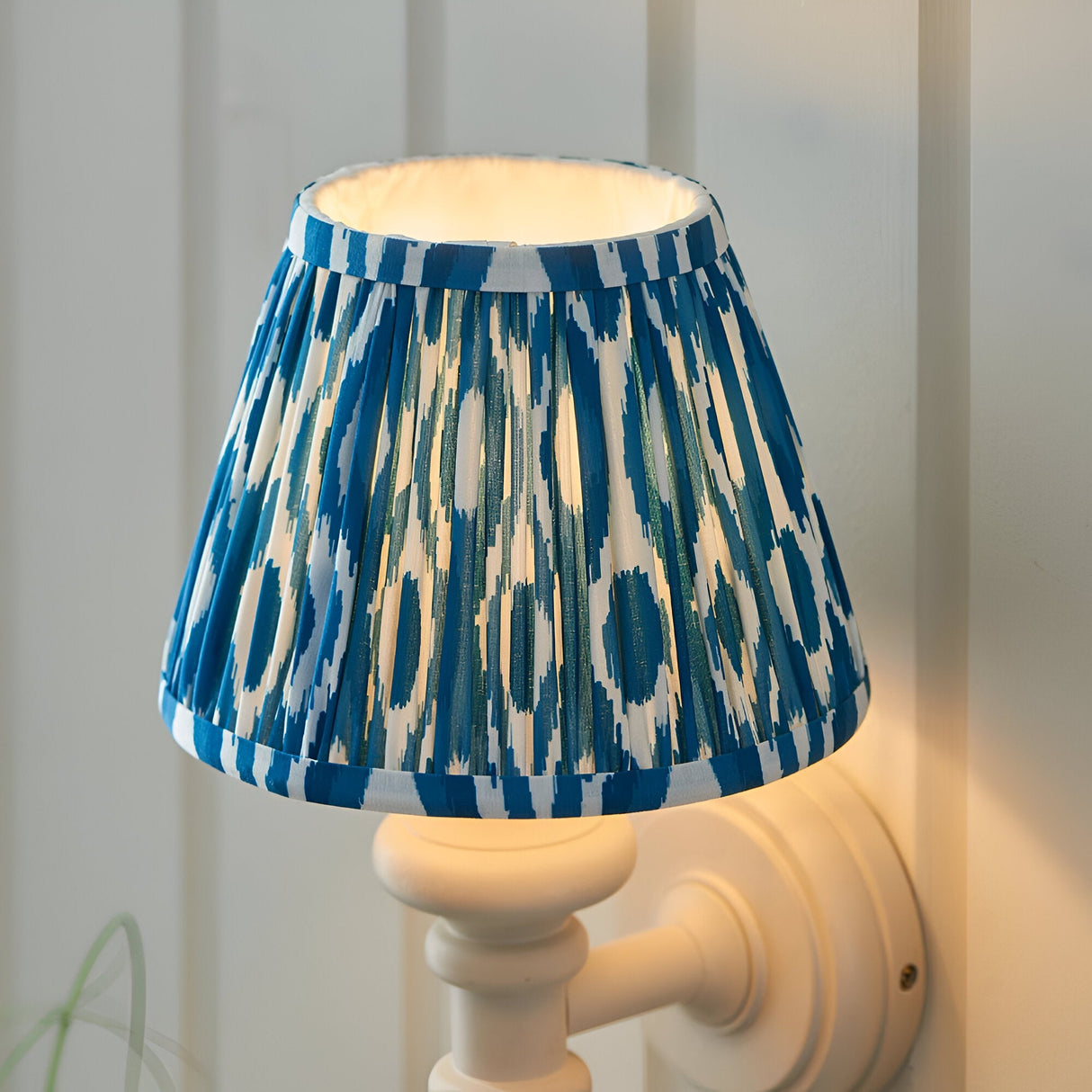 Bevbury Ikat wall light with a pleated blue and white patterned shade on a white turned spindle base, shown illuminated against a panelled wall.