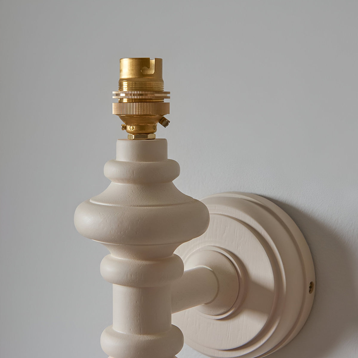 Bevbury Ikat wall light base close-up showing turned white spindle, brass B22 lamp holder, and wall bracket against a light background.