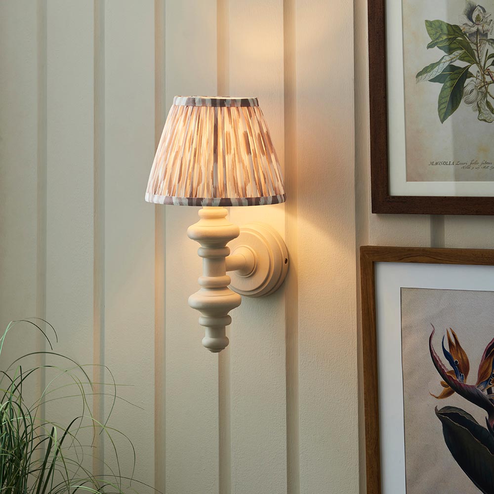 Endon Bevbury Ikat wall light with turned white spindle, brass B22 holder and tapered natural Ikat shade, lit beside framed botanical prints on panelling.