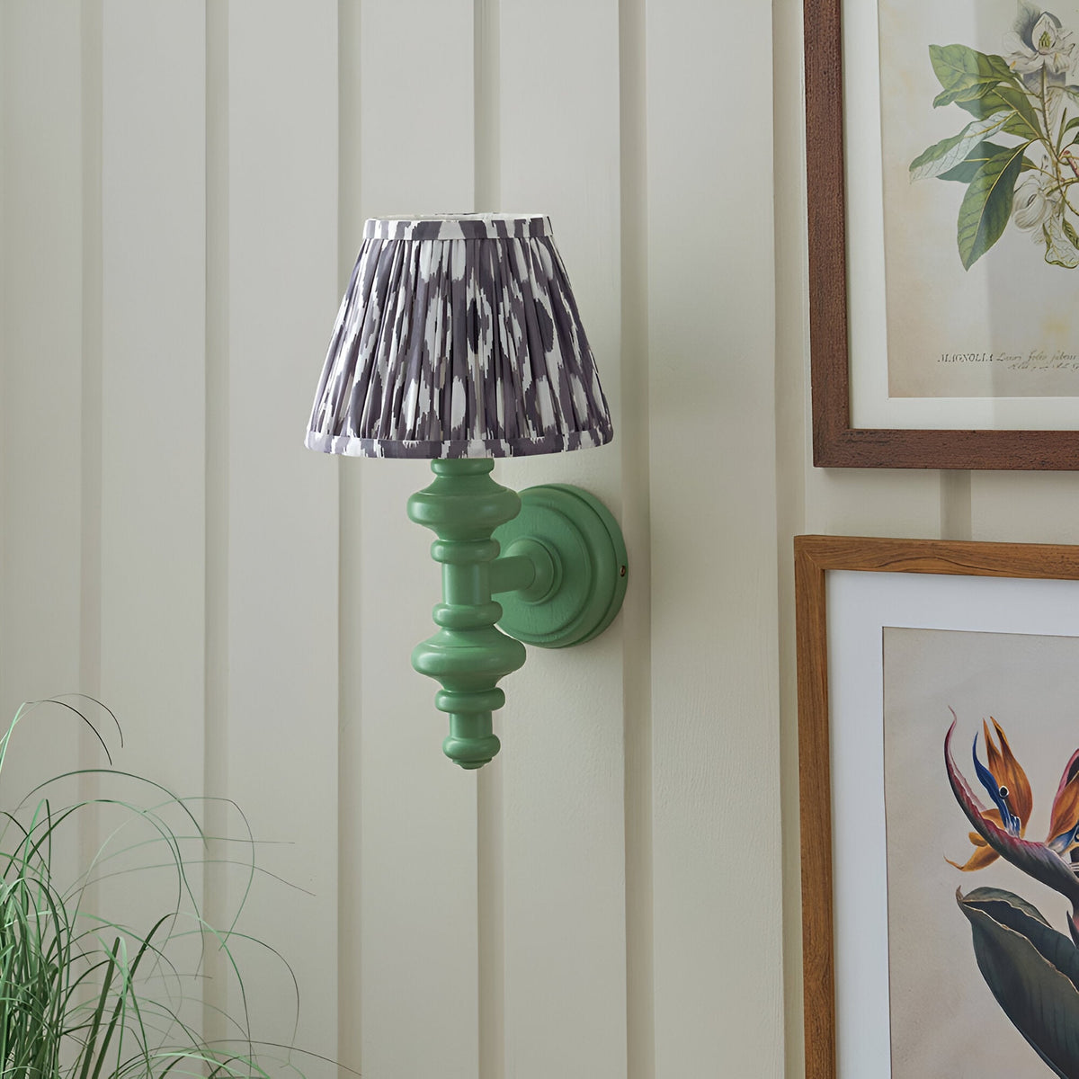 Bevbury Ikat wall light with green turned spindle base, brass holder and grey patterned shade, mounted near framed botanical prints.