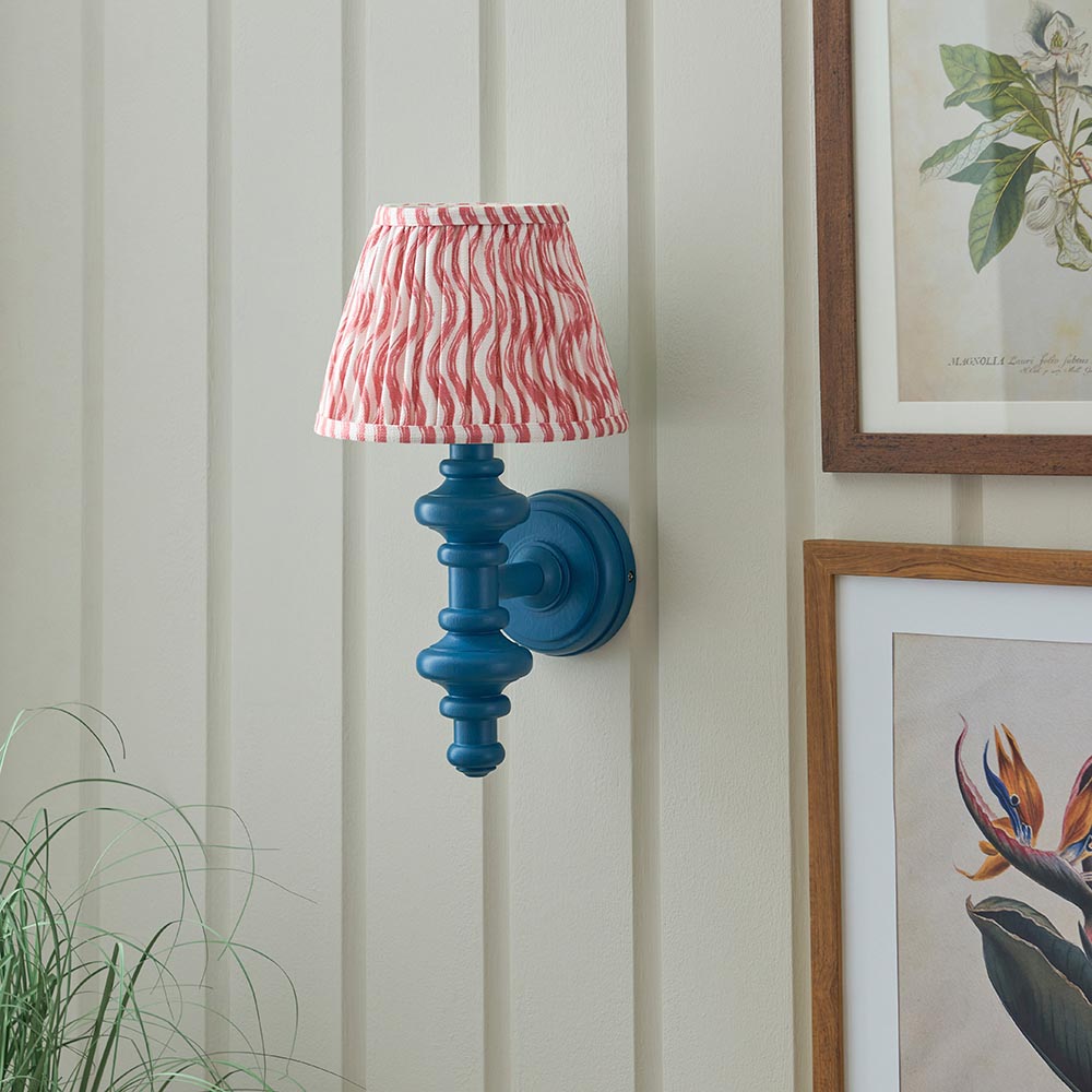 Endon Bevbury Ripple wall light with blue turned wood spindle, brass B22 holder and pink striped faux linen shade, mounted beside framed botanical prints.