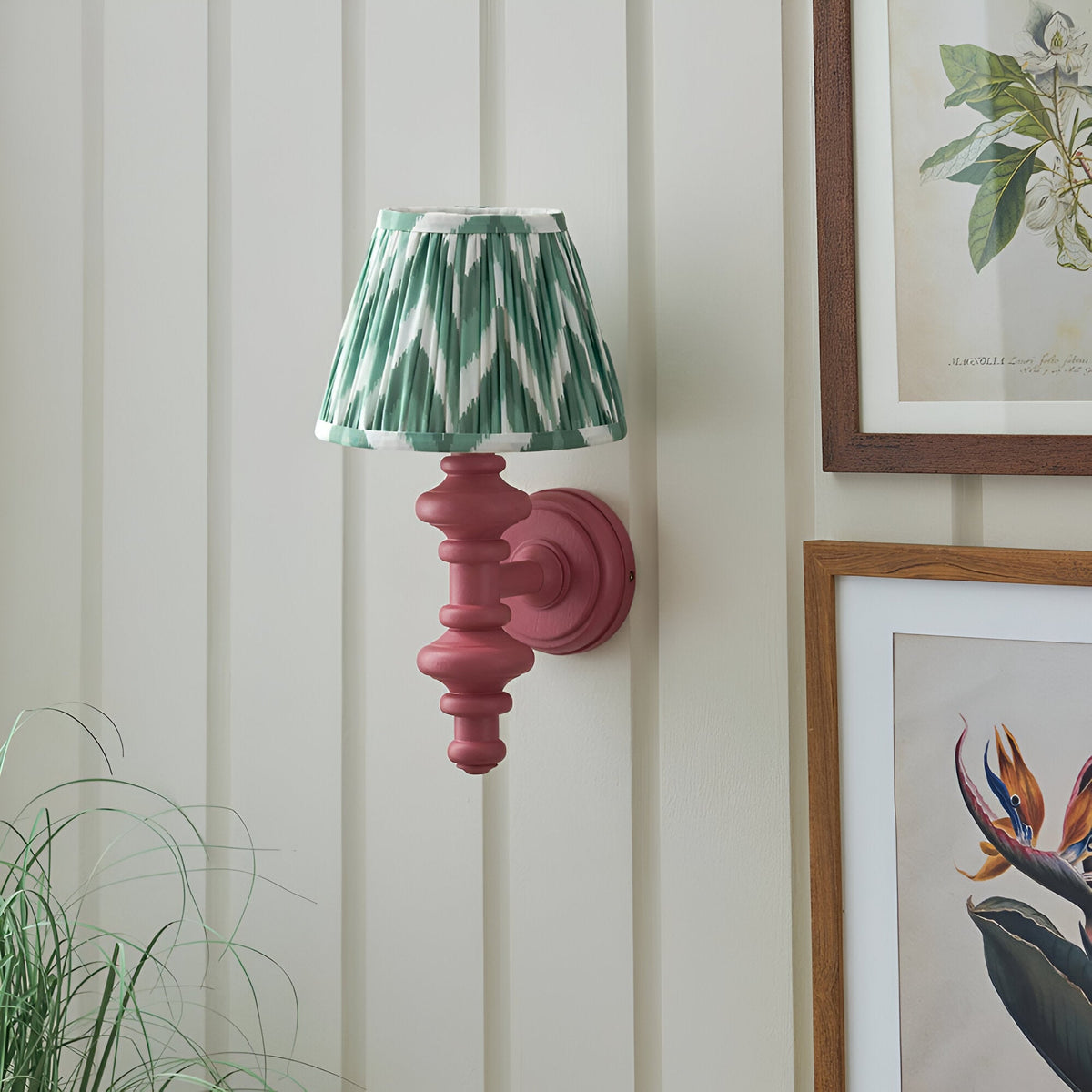 Endon Bevbury Zigzag wall light with a pink turned wooden spindle and pleated green zigzag shade, mounted on white panelling beside framed botanical prints.