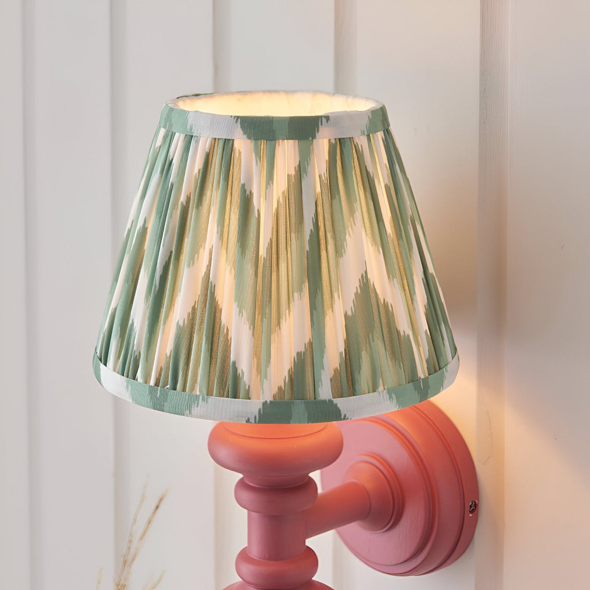 Endon Bevbury Zigzag wall light with pink turned wood spindle, brass B22 holder and green pleated faux silk shade, mounted on white panelling.