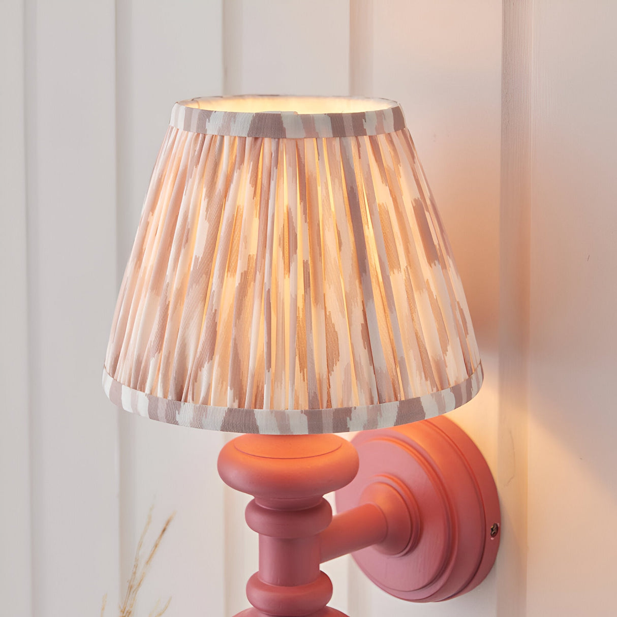 Bevbury Ikat wall light with pink turned wooden spindle base and pleated natural Ikat fabric shade, mounted on a light panelled wall.