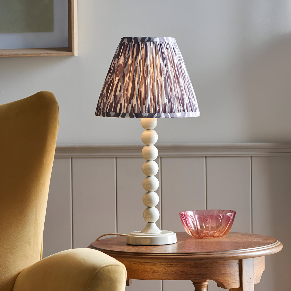 Brenleigh Ikat table lamp with white stacked-sphere metal base and pleated grey and brown Ikat fabric shade, shown on a wooden side table.