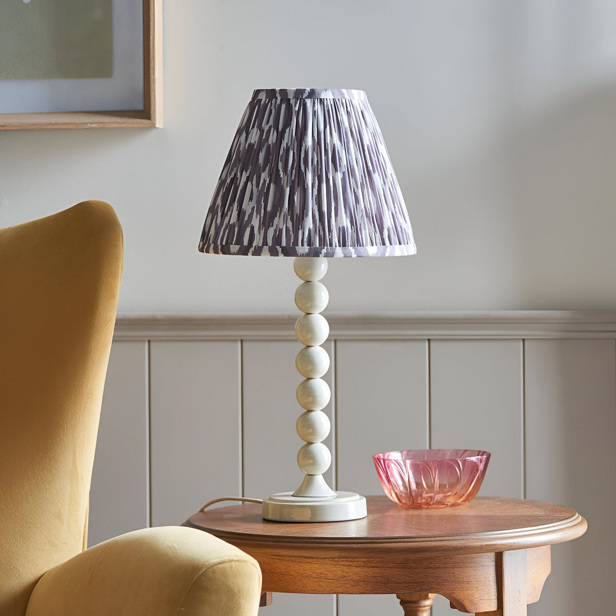 Brenleigh Ikat table lamp with white stacked-sphere base and pleated grey patterned shade, shown on a wooden side table beside a pink glass bowl.