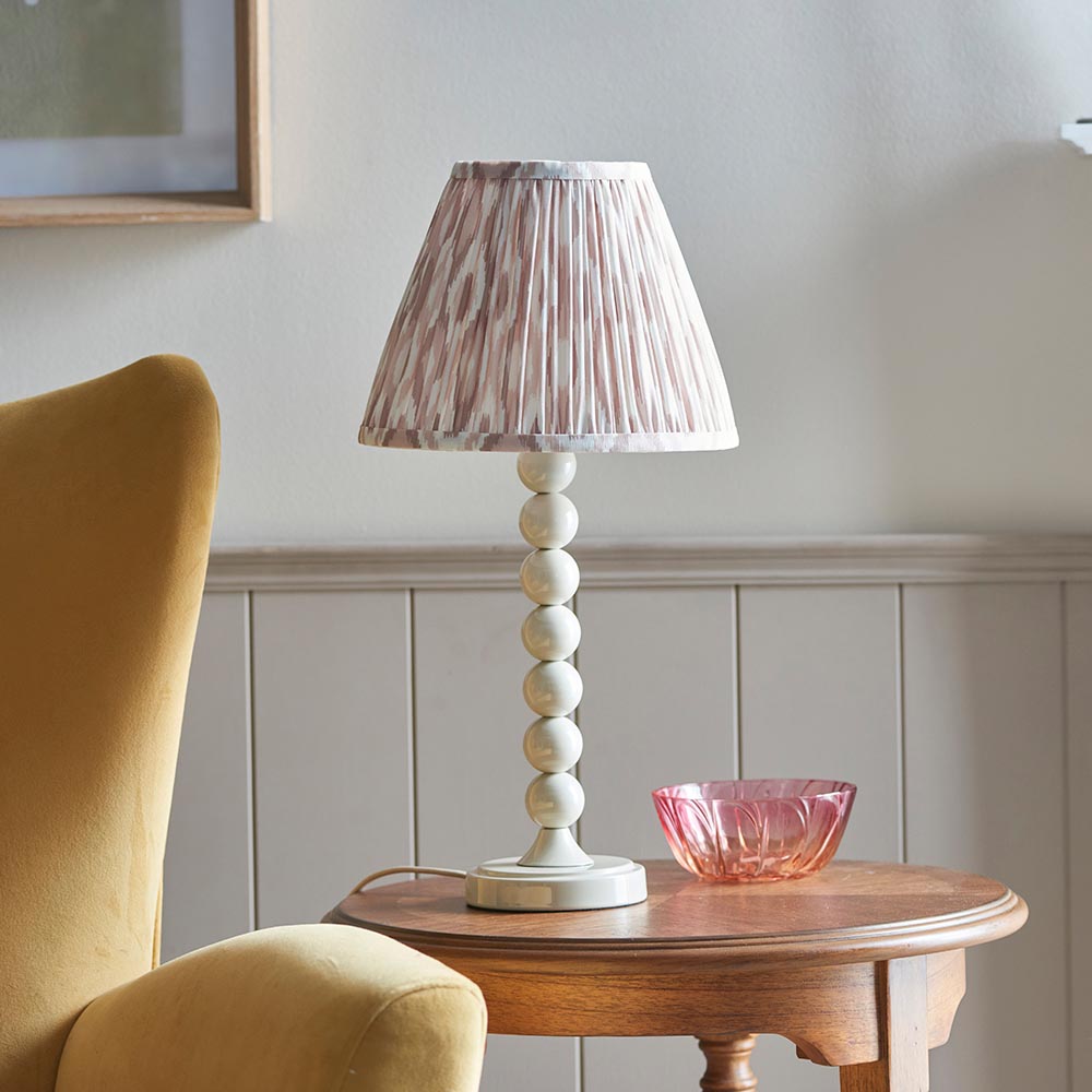 Brenleigh Ikat Table Lamp with stacked-sphere white base and pleated pink-toned Ikat shade, shown on a wooden side table beside a pink glass bowl.