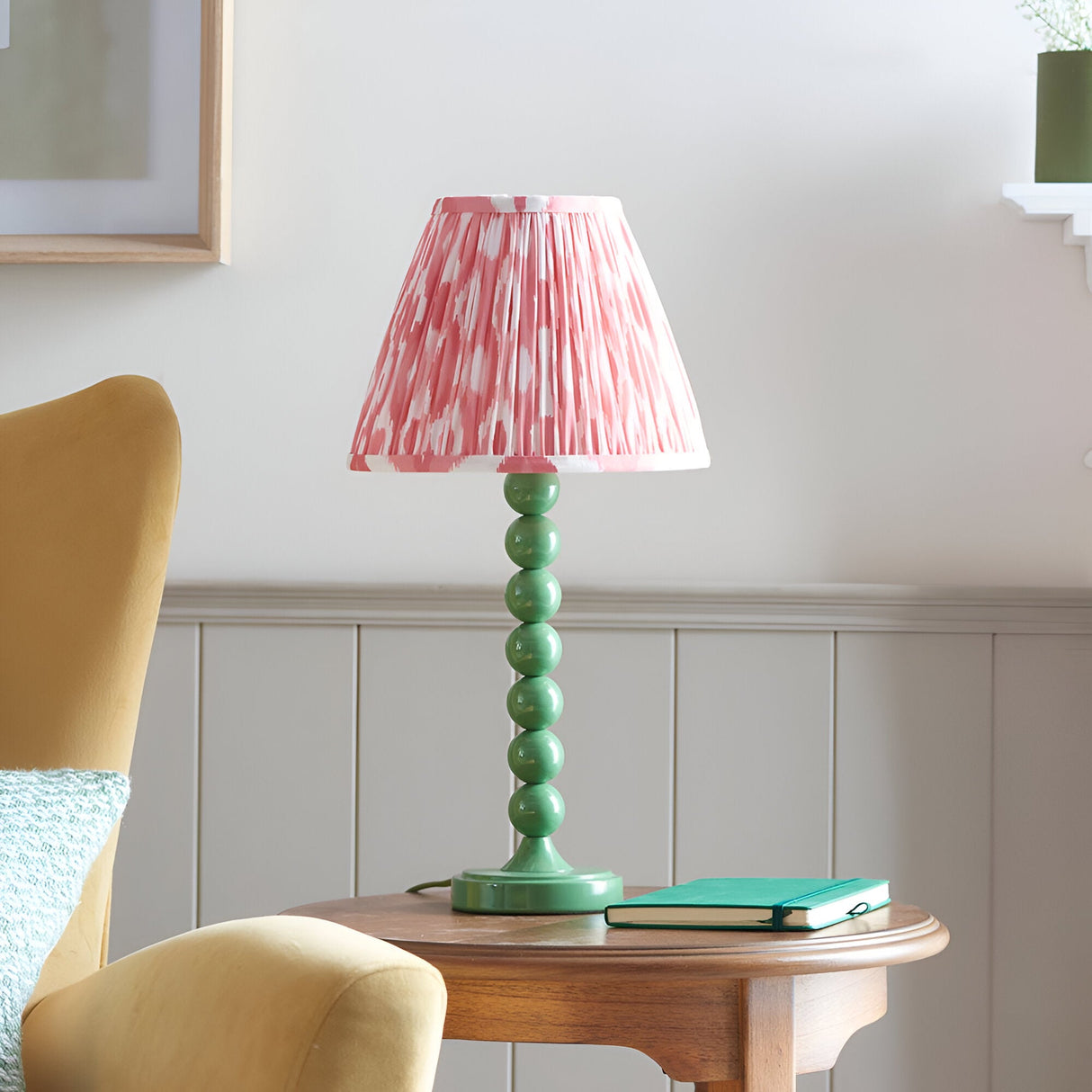 Endon Brenleigh Ikat table lamp with stacked green sphere base and pink ikat pleated shade, on a wooden side table beside a mustard armchair.