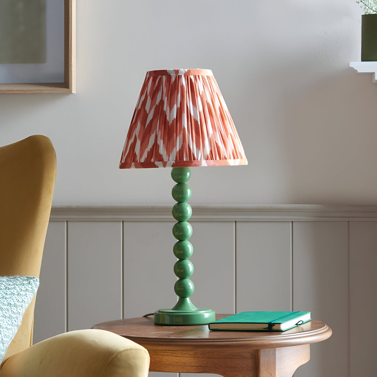 Brenleigh Zigzag Table Lamp in green, with a stacked-sphere metal base and pleated coral zigzag fabric shade, shown on a side table in a living room.