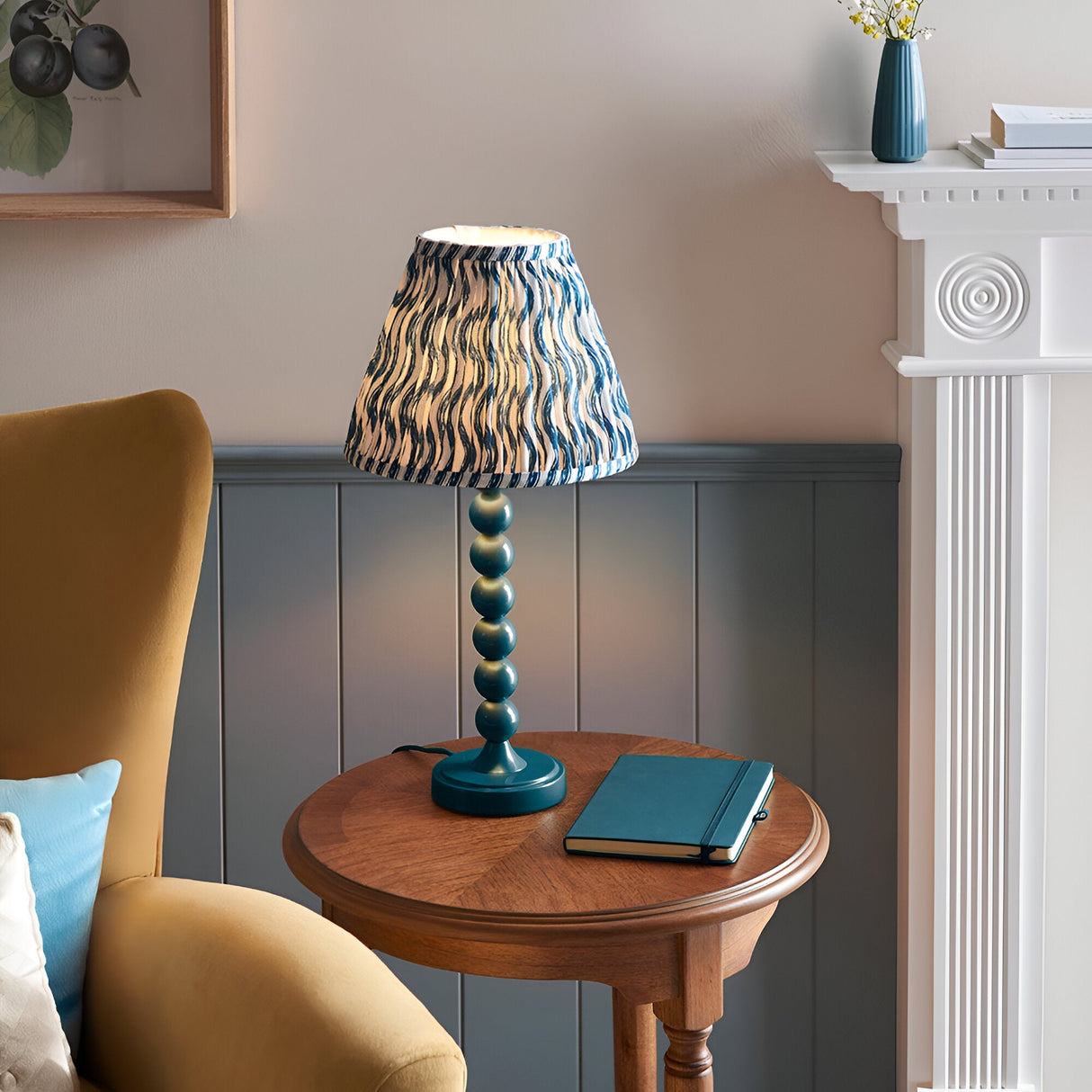 Brenleigh Ripple Table Lamp in blue, with stacked-sphere gloss metal base and pleated wavy-print fabric shade, shown on a side table in a living room.