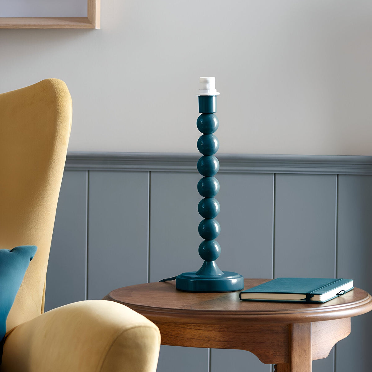 Endon Brenleigh table lamp base in glossy teal with stacked-sphere metal column, shown on a wooden side table beside a yellow armchair and blue notebook.