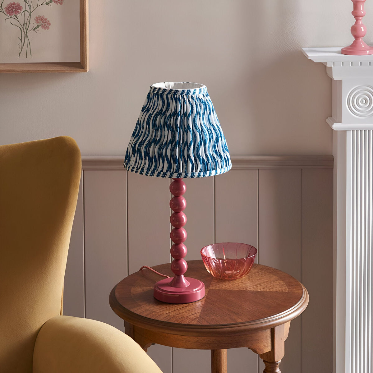 Endon Brenleigh Ripple table lamp with pink stacked-sphere base and blue ripple-print pleated shade on a wooden side table beside a yellow armchair.