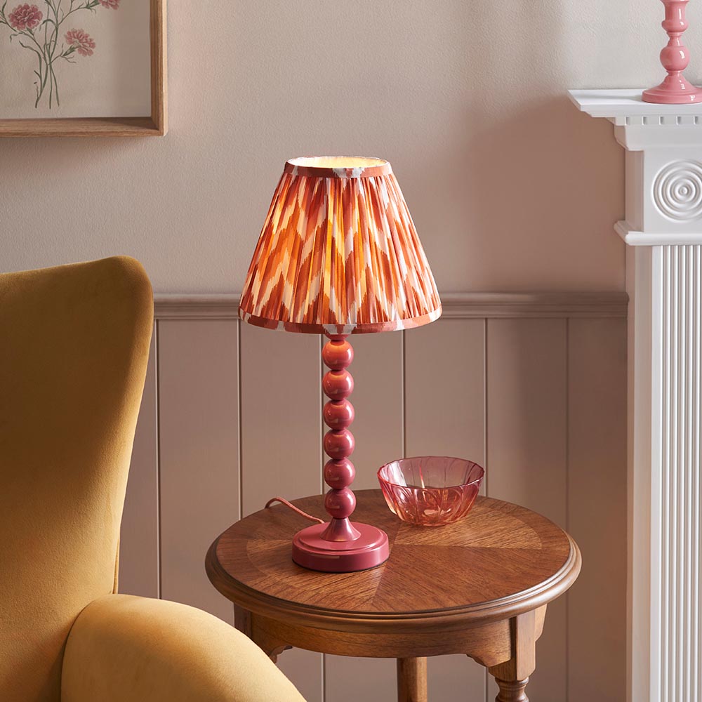 Brenleigh Zigzag Table Lamp in pink with stacked-sphere metal base and pleated zigzag-print shade, shown on a side table in a living room setting.