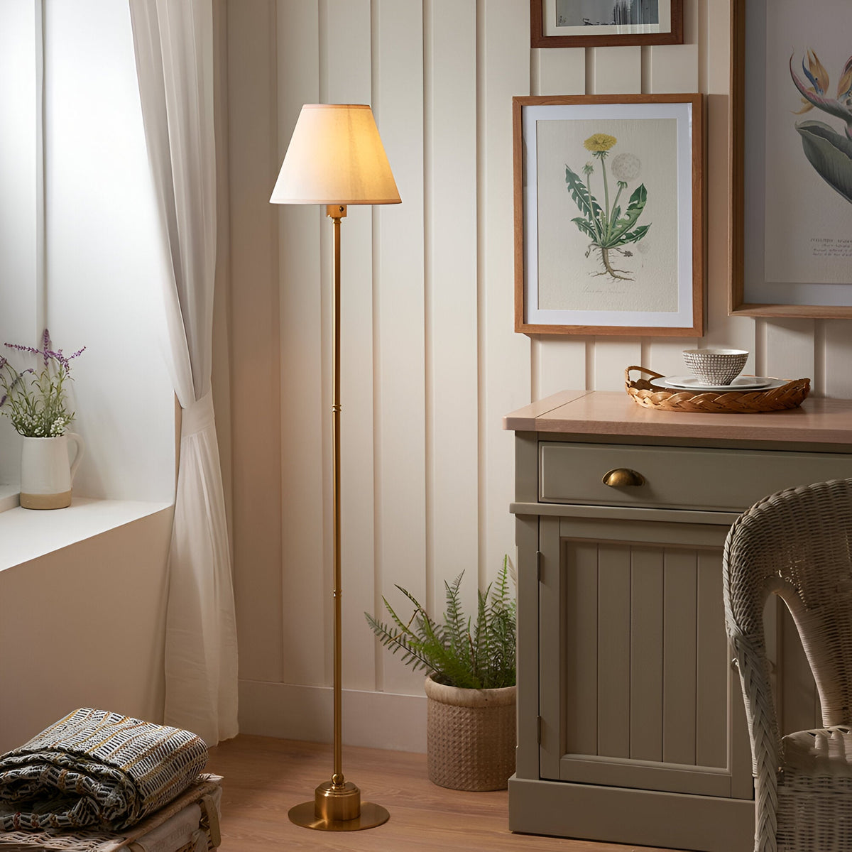 Endon Ashstead LED floor lamp with brushed aged brass stem and vintage white Ivy fabric shade, lit beside a cream panelled wall in a cosy living room.