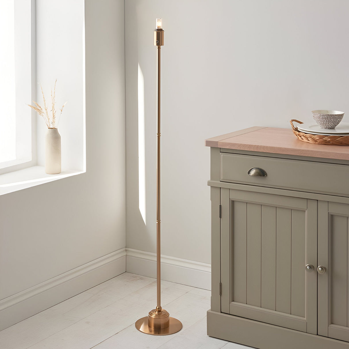 Endon Ashstead LED floor lamp in aged brass without shade, slim stem on a round base, shown beside a sage sideboard in a bright neutral room.