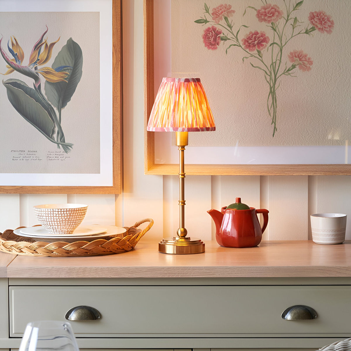 Endon Ashstead LED table lamp with brushed brass stem and pleated pink ikat shade, lit on a wood counter beside botanical prints, a red teapot and dishes.
