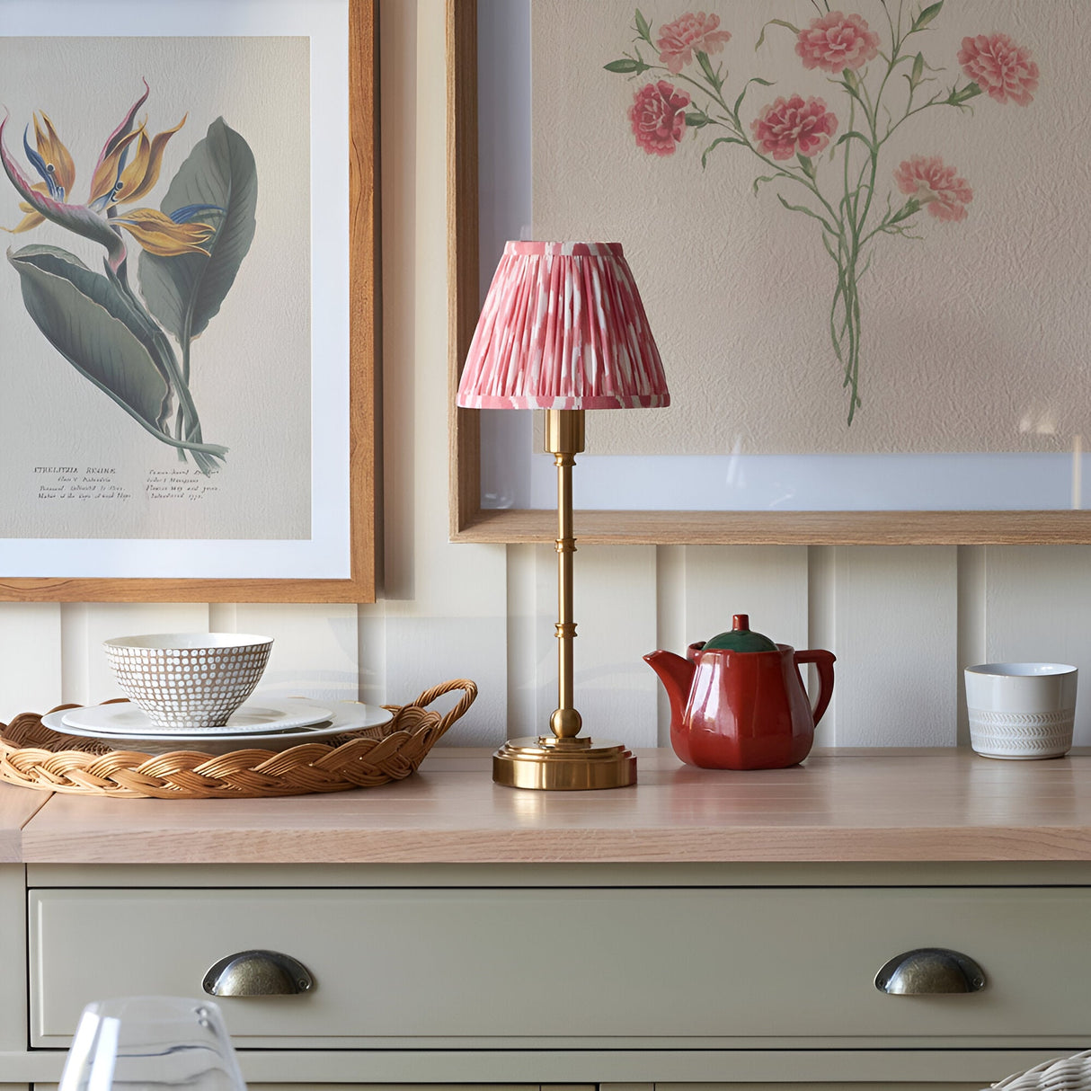 Endon Ashstead LED table lamp with brushed brass stem and pleated pink ikat shade, on a wood countertop with botanical prints and ceramics.
