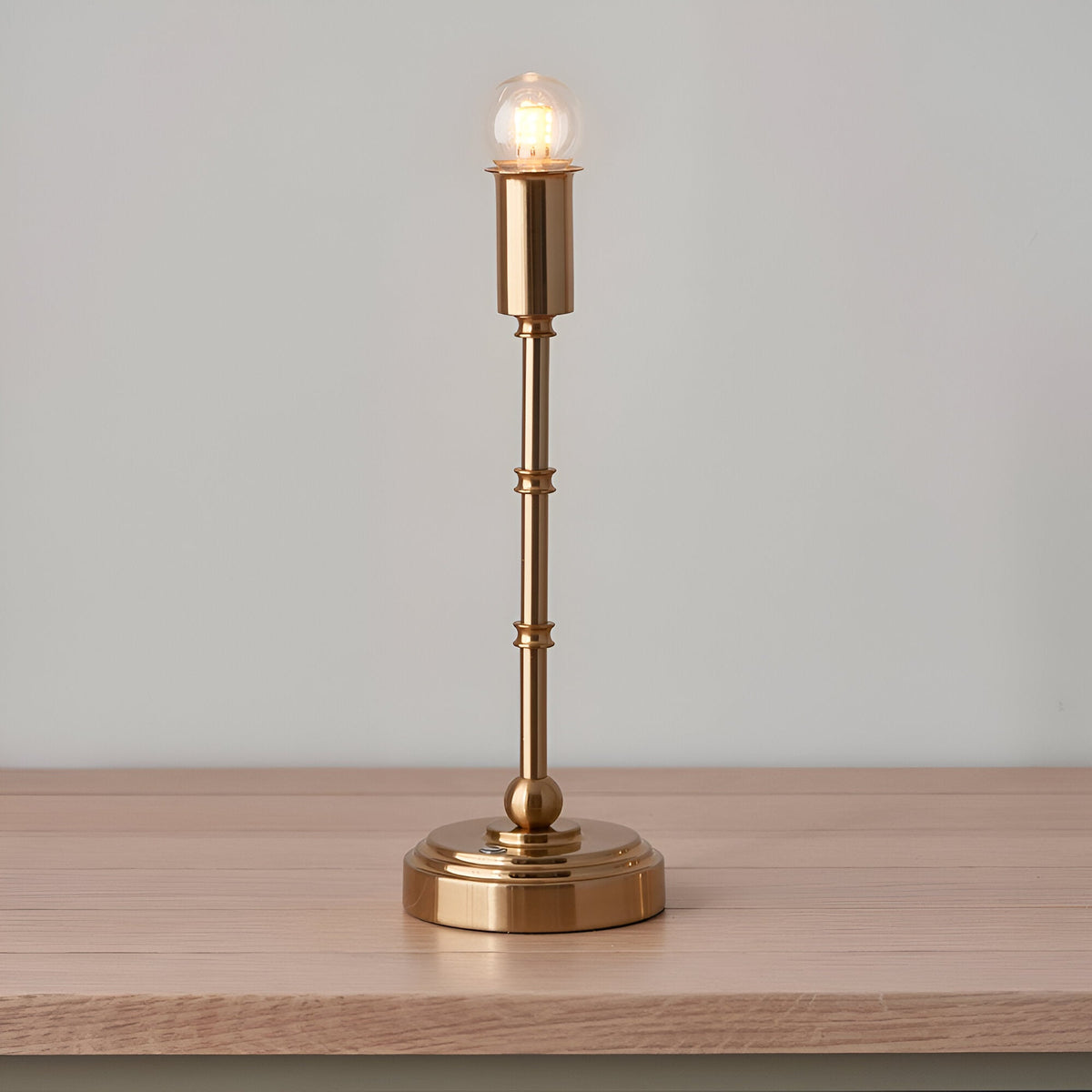 Endon Ashstead cordless table lamp in brushed brass with exposed LED bulb, shown without shade on a light wood surface against a pale background.