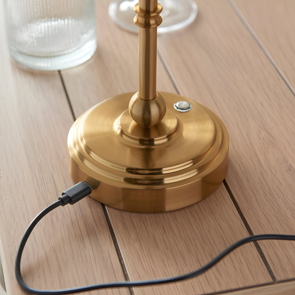 Close-up of the Endon Ashstead table lamp's brushed brass base with USB-C charging cable connected, shown on a light wooden slatted surface.