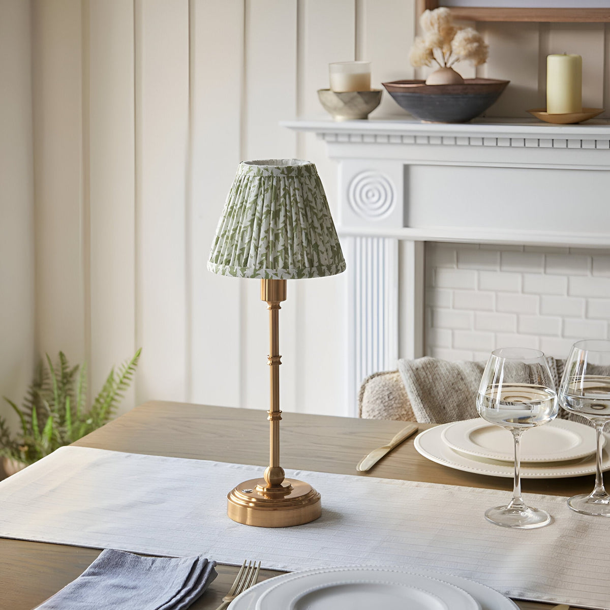 Ashstead LED Leaf Table Lamp in brass with a pleated green and white leaf-print shade, styled on a dining table near a white fireplace.