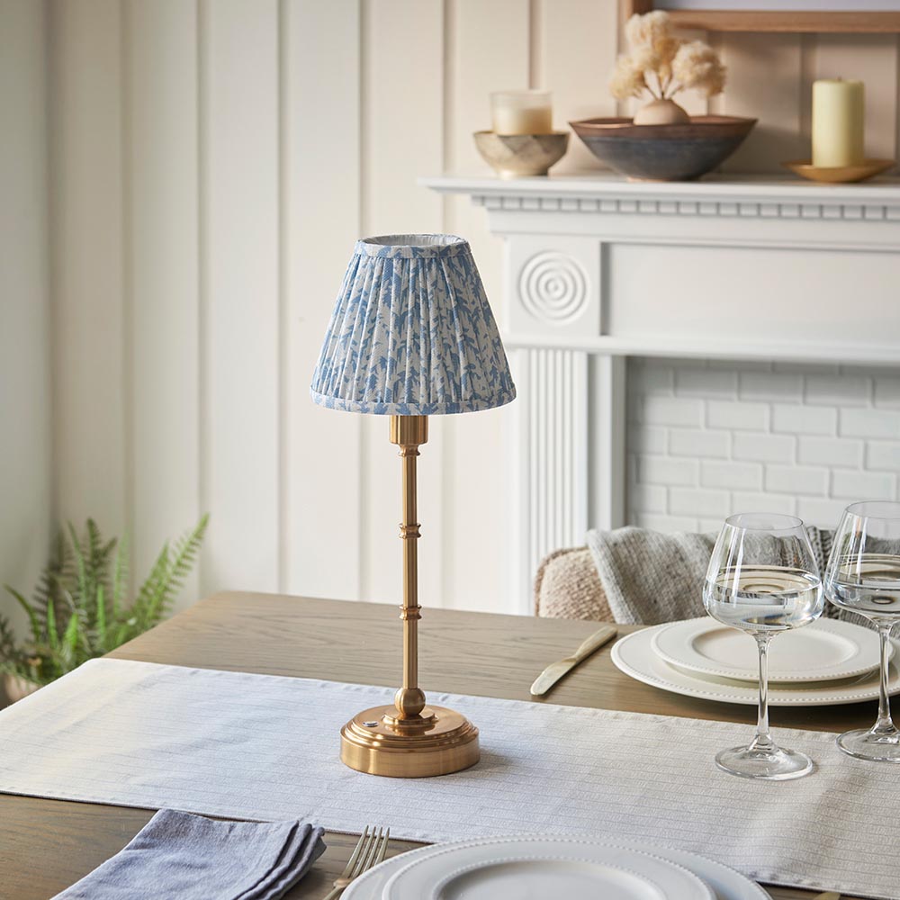 Ashstead LED Leaf Table Lamp with brass base and pleated blue leaf-print fabric shade, shown on a dining table beside white plates and glassware.