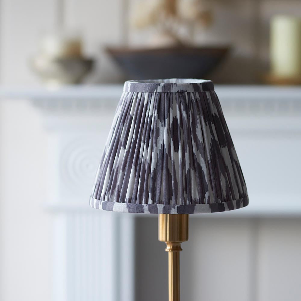 Ashstead LED table lamp with pleated grey and white Ikat fabric shade on a brass base, shown on a mantelpiece in a living room setting.