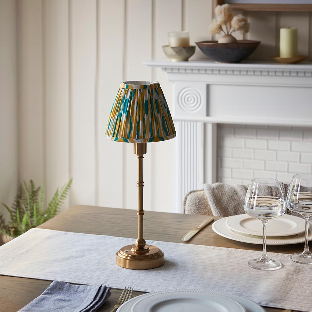 Ashstead LED table lamp with brass stem and base, topped by a yellow, teal and white Ikat fabric shade, shown on a dining table near a fireplace.