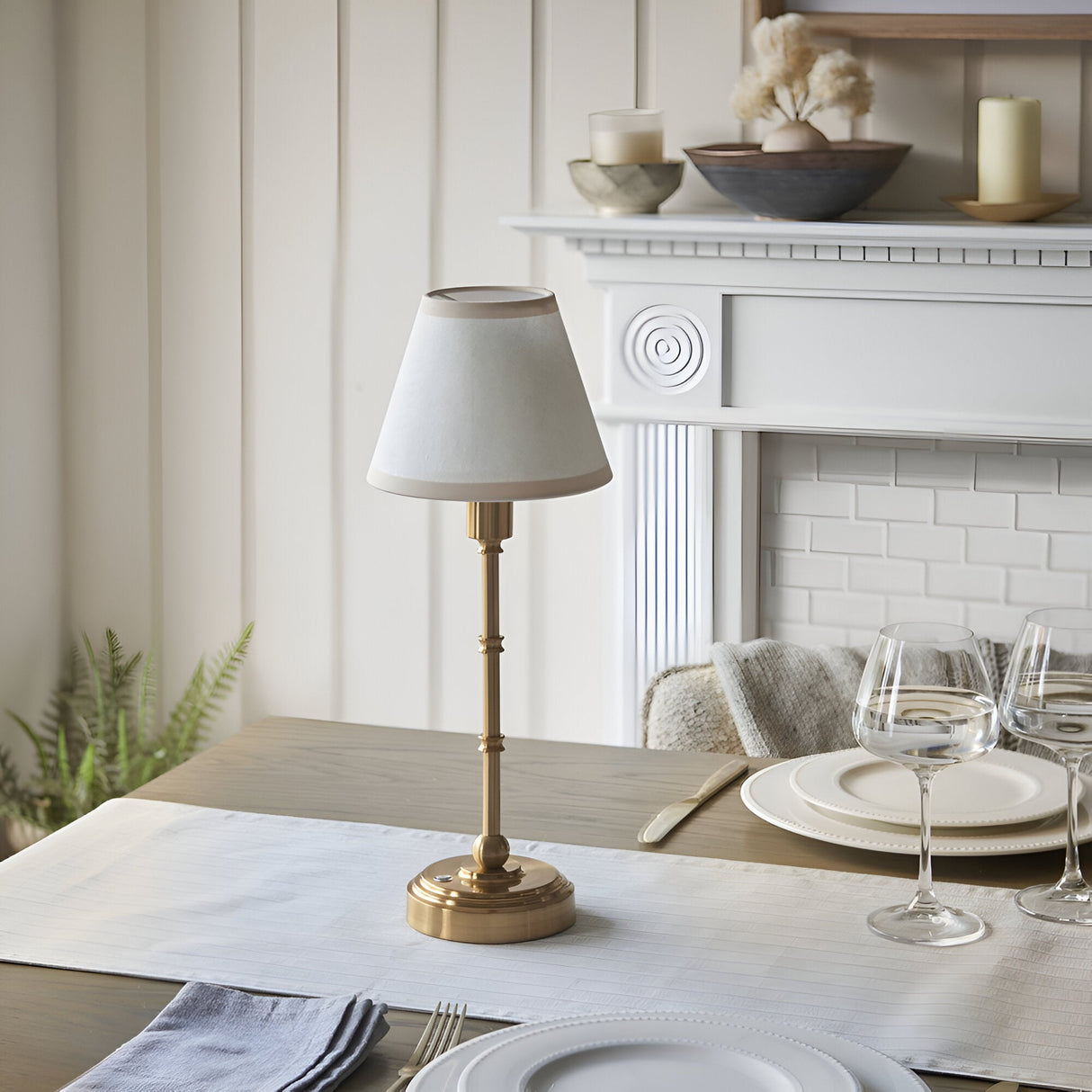 Ashstead LED Table Lamp in brushed aged brass with vintage white fabric shade, styled on a dining table beside a white mantelpiece.