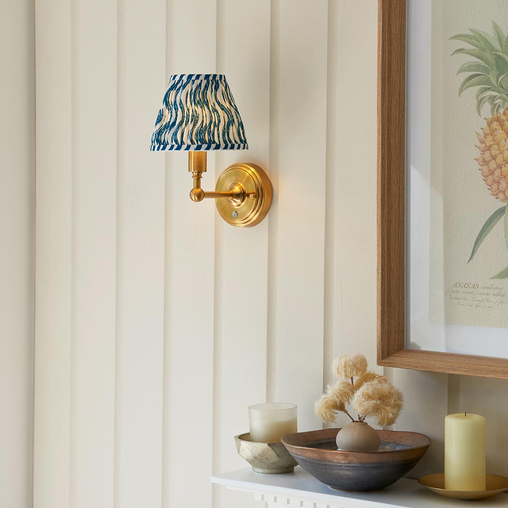 Ashstead LED Ripple wall light with brushed brass arm and blue-and-white patterned fabric shade, mounted beside framed pineapple artwork.