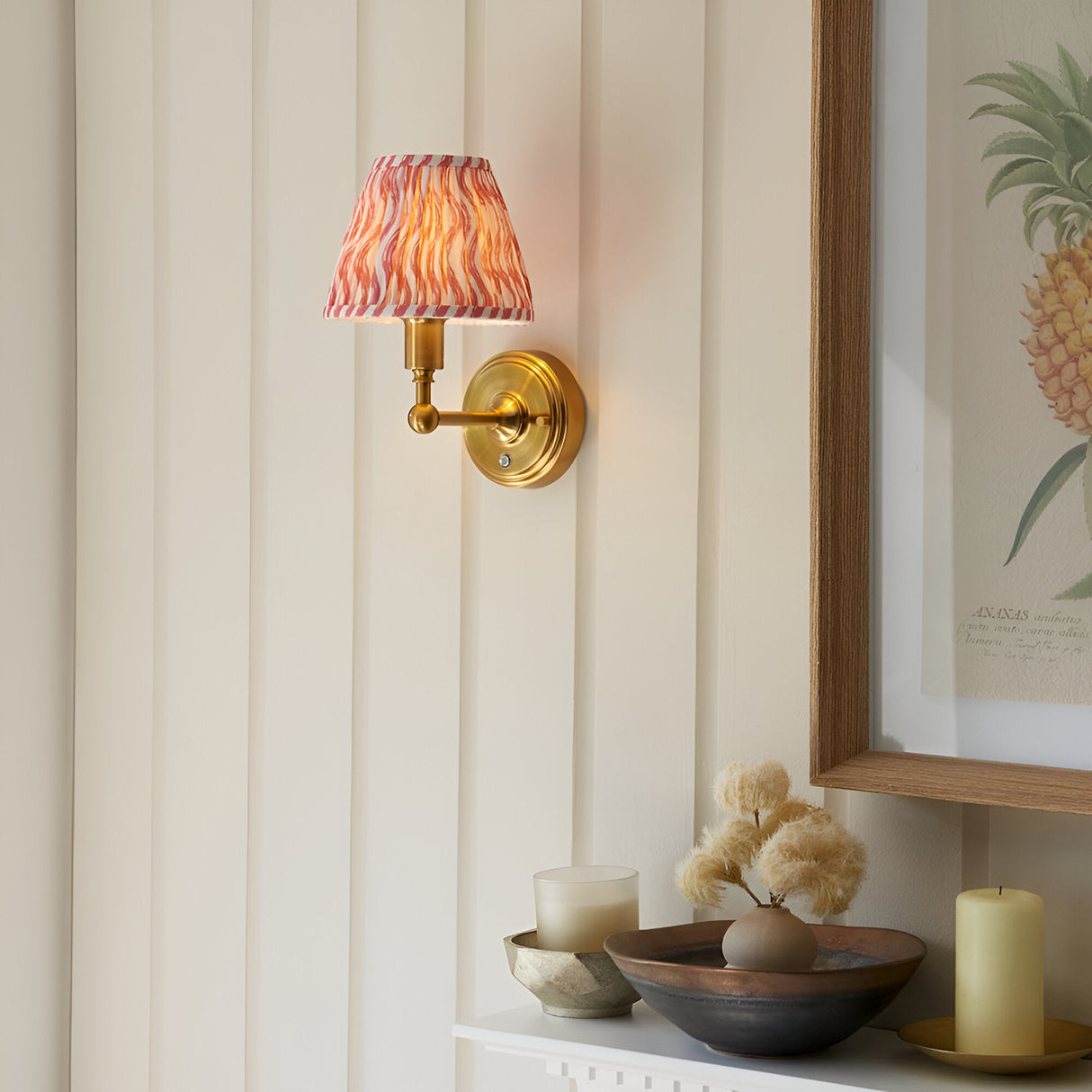 Endon Ashstead LED wall light with brushed brass arm and pink ripple-print shade on a cream panelled wall beside a framed pineapple print.