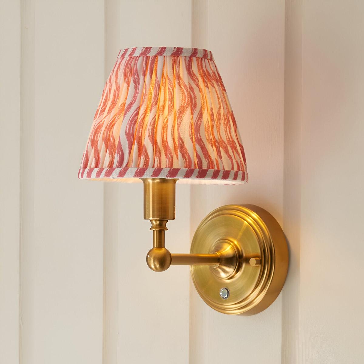 Endon Ashstead LED Ripple wall light with brushed brass arm and pleated coral pink and white patterned fabric shade, mounted on a cream panelled wall.