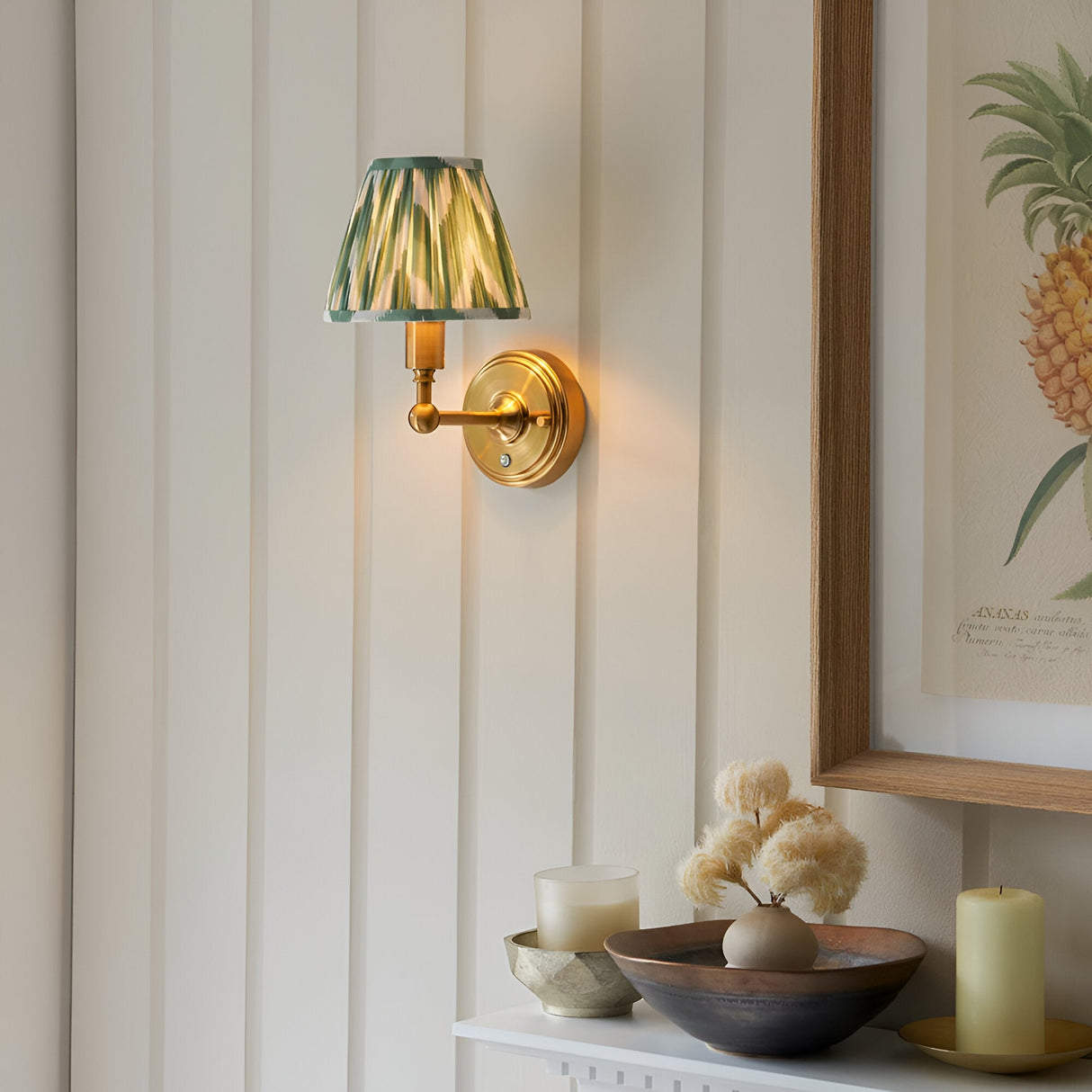 Endon Ashstead LED wall light in brushed brass with green and cream zigzag pleated shade, mounted on panelled wall beside framed pineapple print.