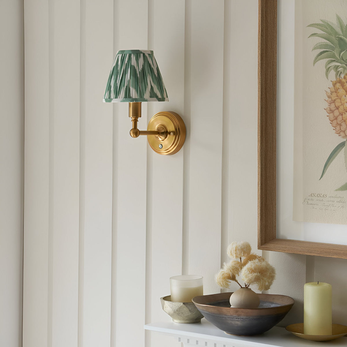 Endon Ashstead LED wall light with brushed brass arm and pleated green zigzag fabric shade, mounted on a panelled wall beside a framed pineapple print.