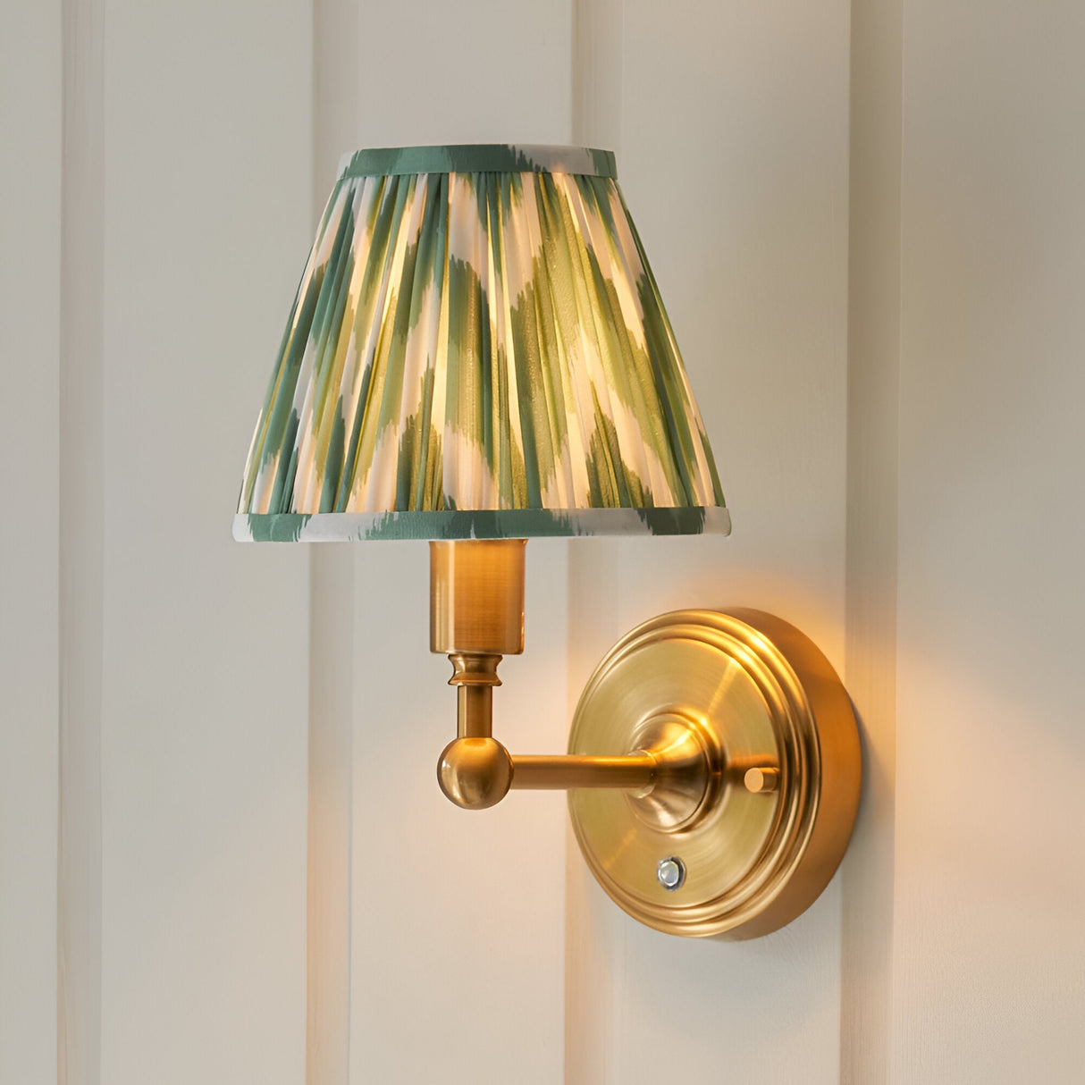 Endon Ashstead LED wall light with brushed aged brass arm and pleated green and white zigzag fabric shade, mounted on a panelled cream wall.