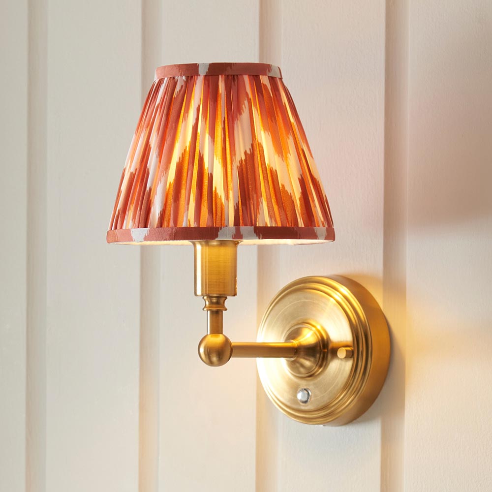 Ashstead LED wall light with orange and cream zigzag-print pleated shade, brushed aged brass arm and circular wall mount, on a panelled white wall.