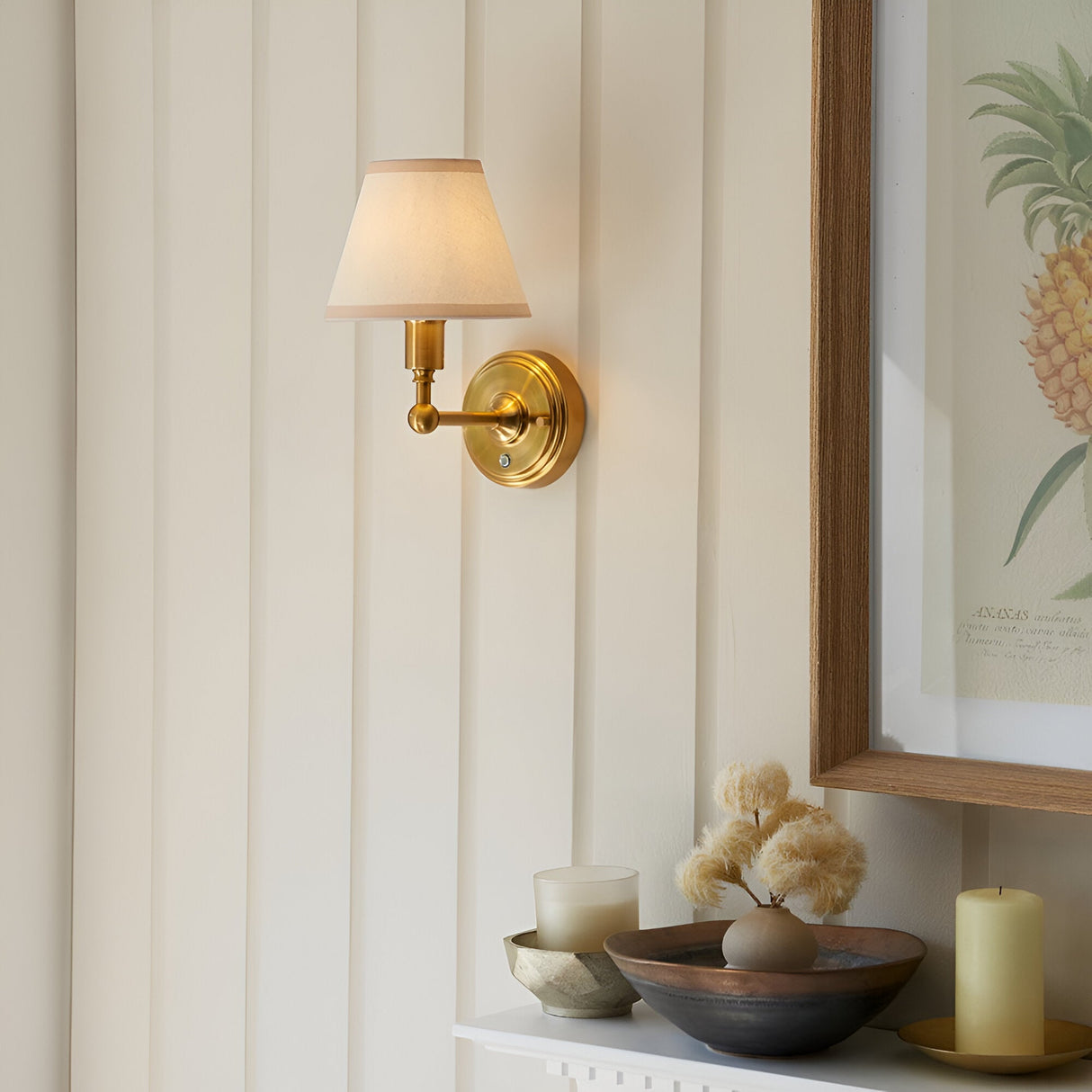 Endon Ashstead LED rechargeable wall light in brushed aged brass with tapered ivory fabric shade, mounted on panelled wall beside framed pineapple print.