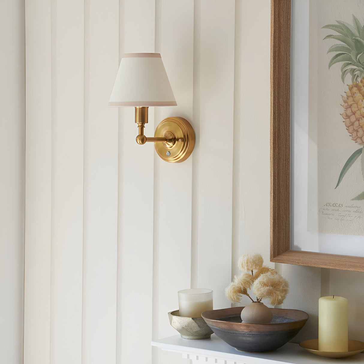 Endon Ashstead LED rechargeable wall light in brushed aged brass with tapered ivory fabric shade, mounted on panelled wall beside a framed pineapple print.