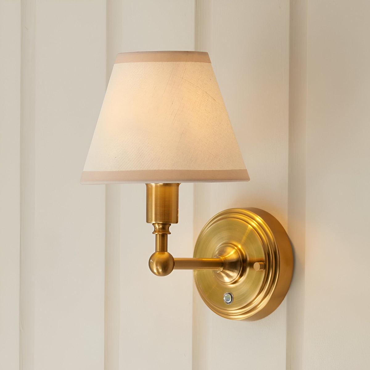 Endon Ashstead LED rechargeable wall light in brushed aged brass with a tapered ivory fabric shade, mounted on a cream panelled wall.