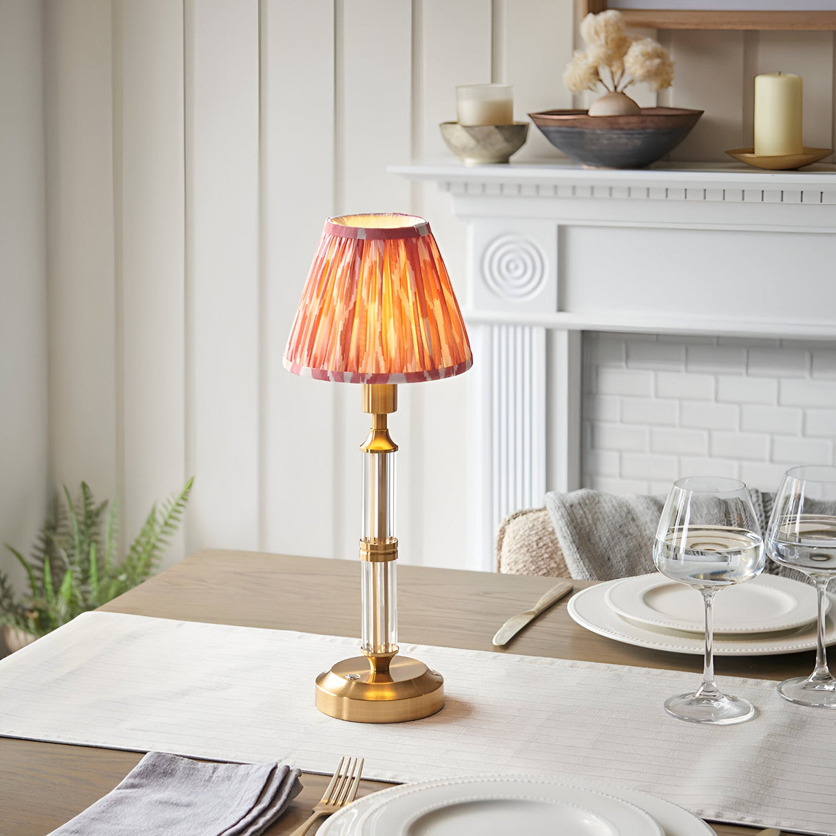 Endon Carlwood LED table lamp with brushed brass base, clear acrylic stem and pleated coral pink Ikat shade, lit on a dining table by a white fireplace.