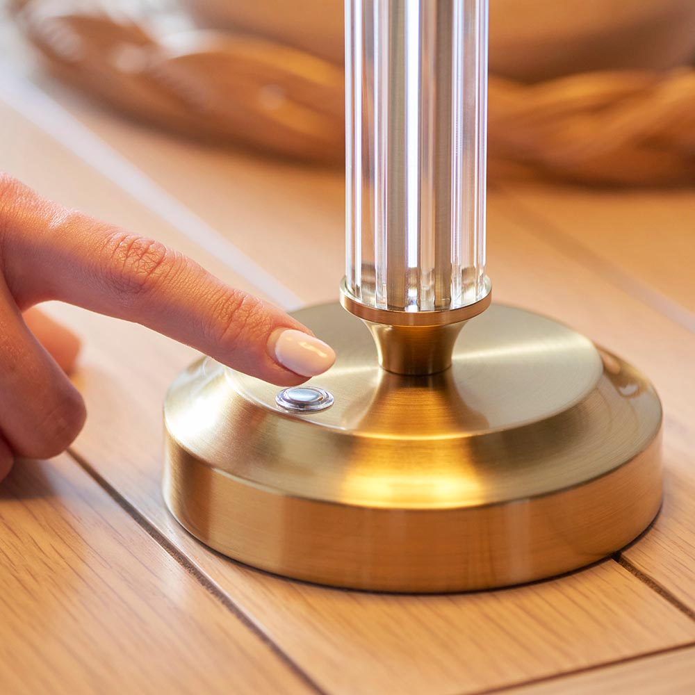 Close-up of the Carlwood LED table lamp's brass base with touch dimmer button, fluted glass column, and finger pressing to switch on.