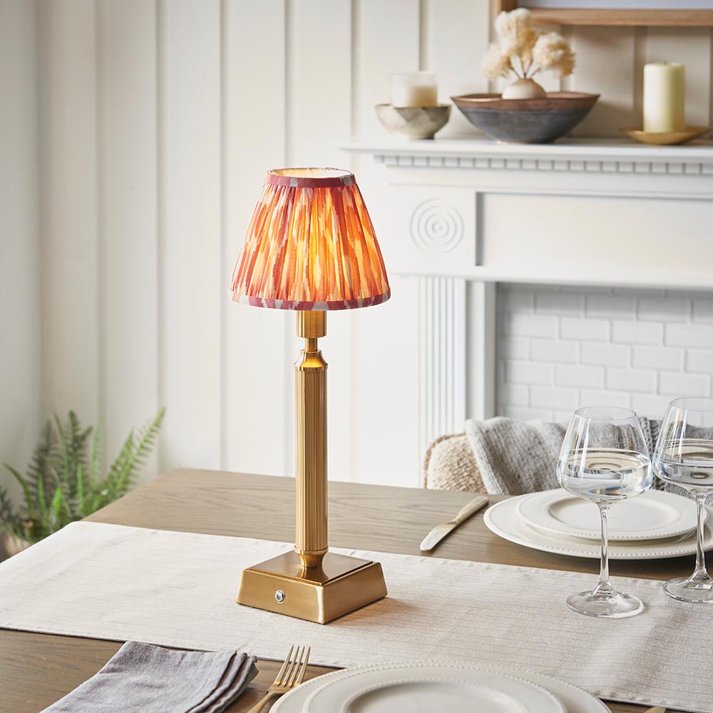 Endon Cranwick LED Ikat table lamp with brushed brass reeded stem, square base and pink and white pleated shade, on a dining table by a fireplace.