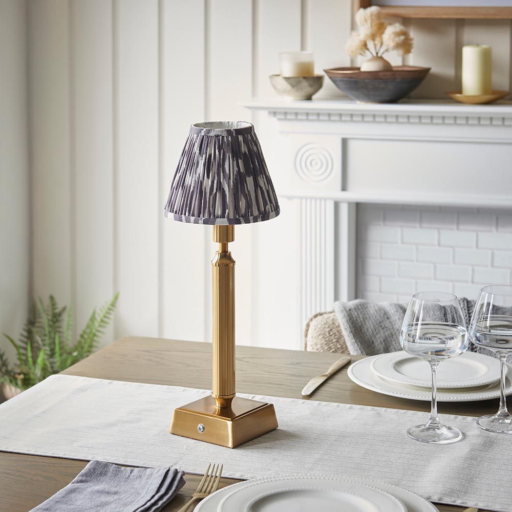 Endon Cranwick LED Ikat table lamp with brushed brass reeded stem and grey and white pleated shade, styled on a dining table set with glassware.