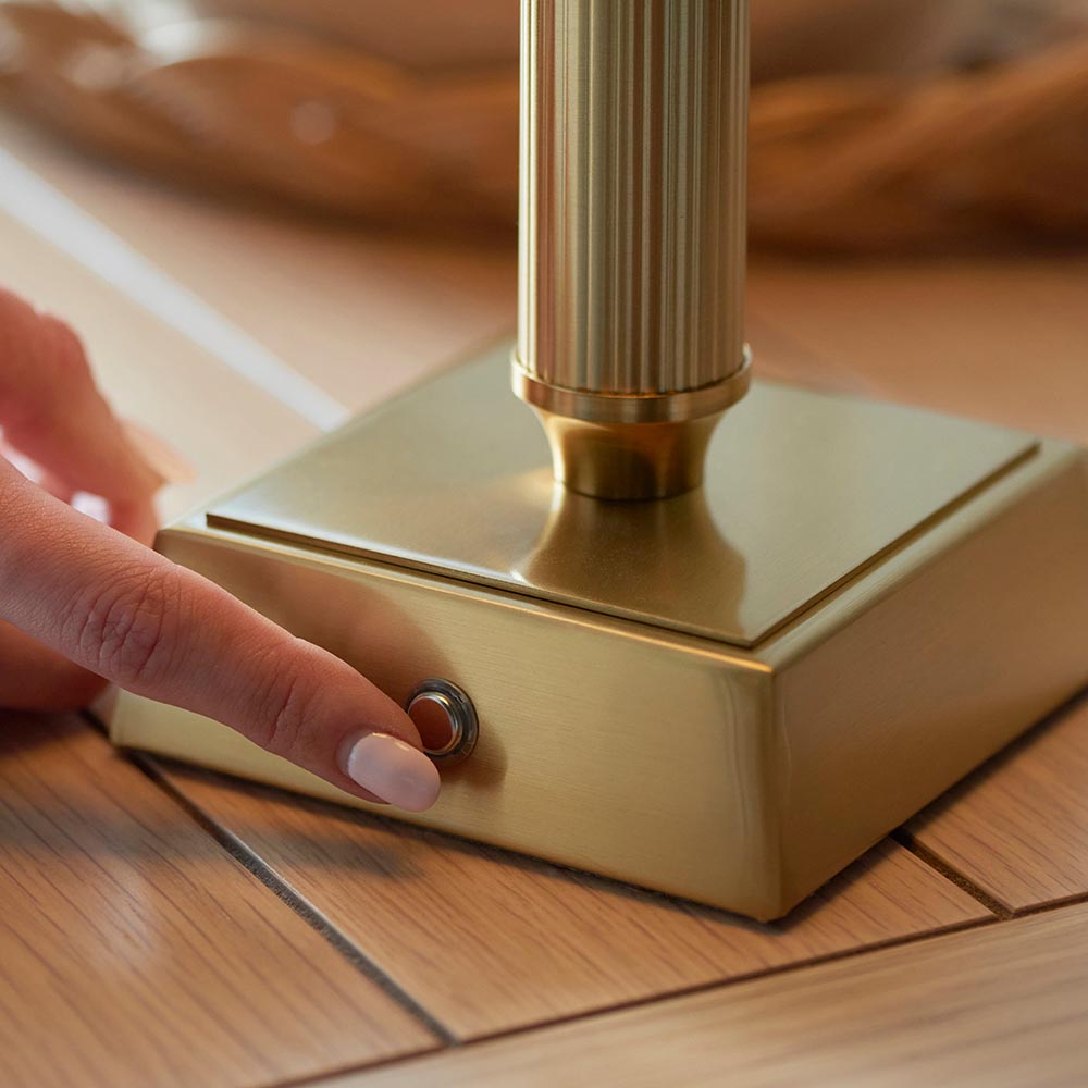 Close-up of a finger pressing the touch-dim button on the brushed brass square base of the Cranwick LED table lamp on a wooden surface.