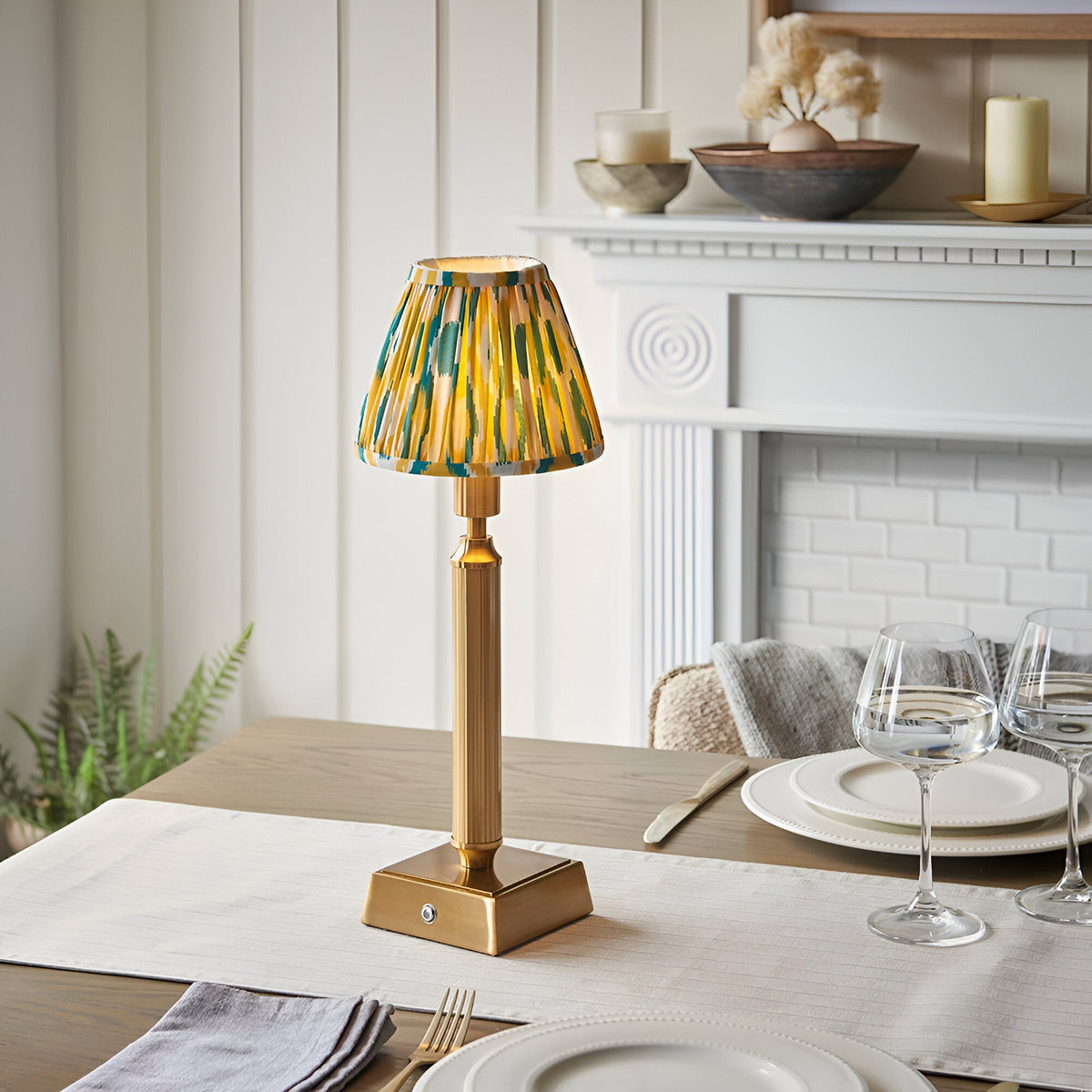 Cranwick LED Ikat Table Lamp with reeded brass stem, square base, and yellow-green patterned fabric shade, shown on a dining table near a fireplace.