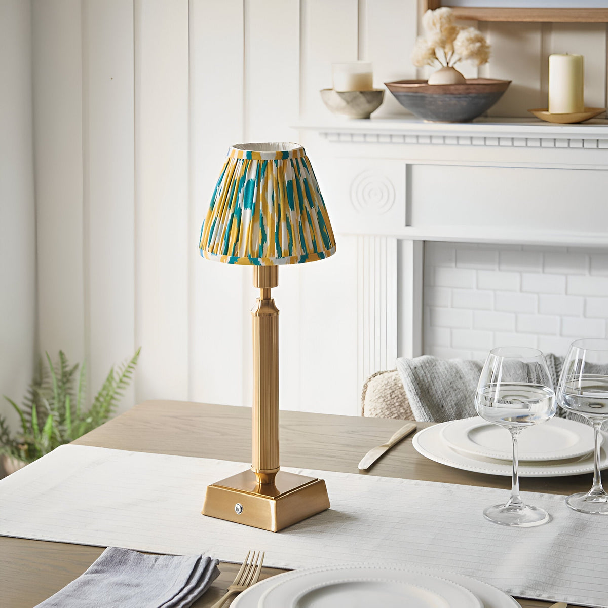 Cranwick LED Ikat table lamp with brass reeded stem, square base, and patterned yellow, teal and white fabric shade, shown on a dining table.