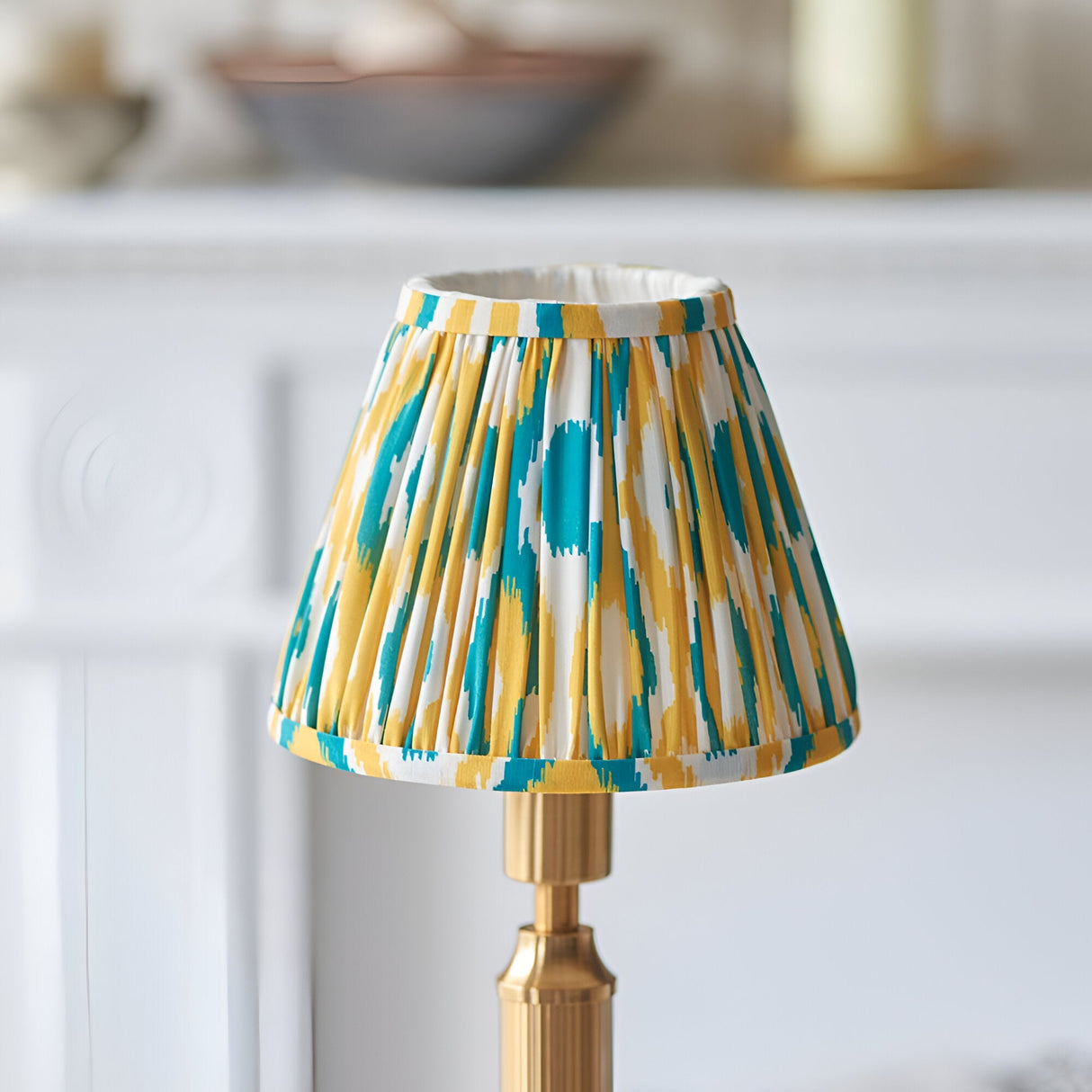 Cranwick LED table lamp with brass reeded stem and pleated Ikat fabric shade in teal, yellow and white, shown in a bright room setting.
