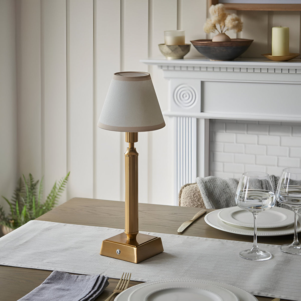 Endon Cranwick rechargeable table lamp with reeded brushed brass stem and vintage white Ivy fabric shade on a dining table beside a white fireplace.