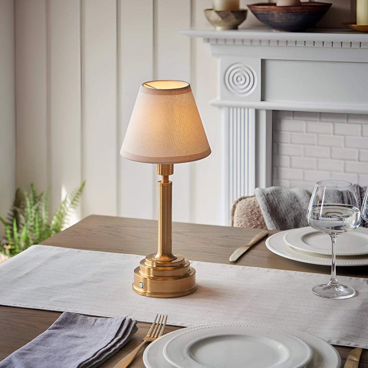 Danebourne LED rechargeable table lamp with brass stepped base and fabric shade, shown lit on a dining table beside a white fireplace.