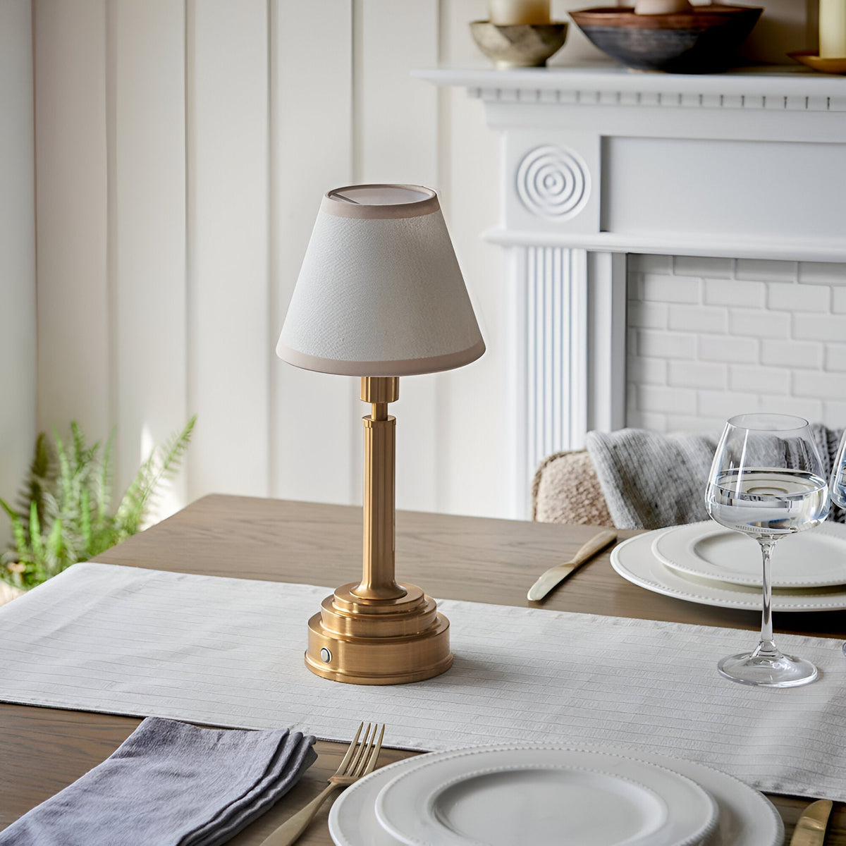 Danebourne LED rechargeable table lamp in brass with cream fabric shade, styled on a dining table beside white plates and a glass, near a white fireplace.