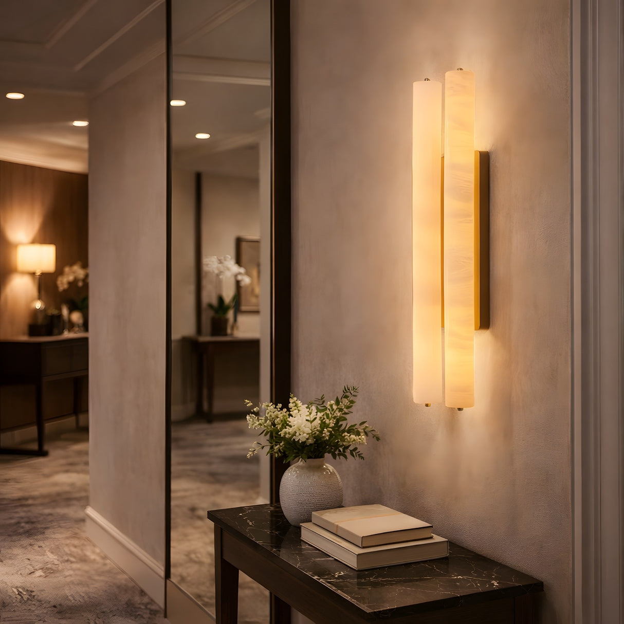 A modern hallway features the Selvara LED Medium Alabaster Double Wall Light in brushed brass and soft white, illuminating a marble-top console table with flowers, stacked books, and a large mirror reflecting the gentle glow.